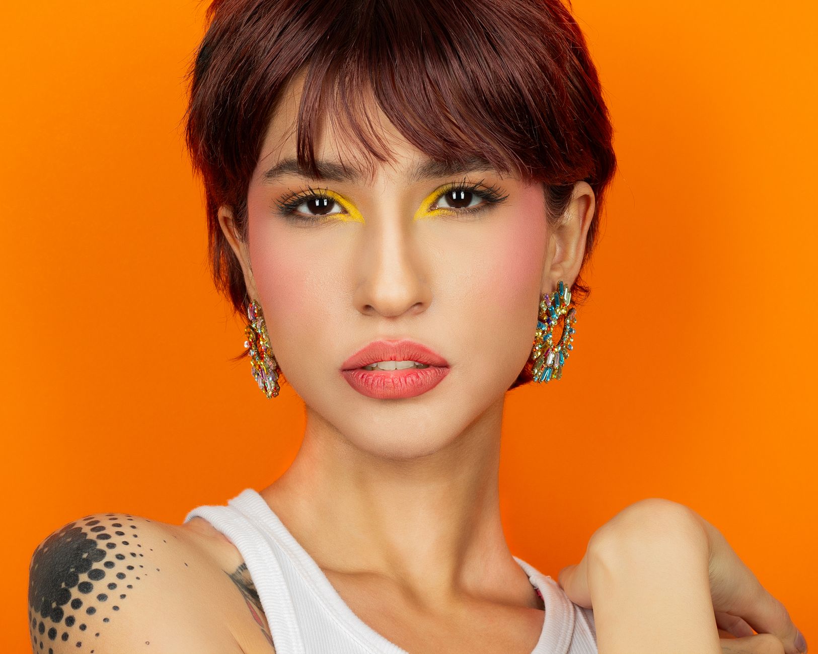 Colorful portrait of a woman with striking makeup and earrings on an orange background. — Why Is Sunscreen Vital To Your Skin